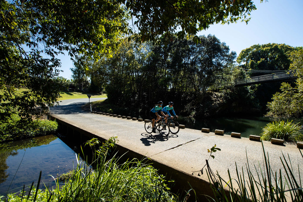 Two triathletes cycling across the river wearing the SYKL PRO cycling jersey